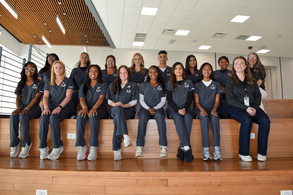 A group of students in uniforms pose for a photo on a stage. Some sit on the edge, others stand.