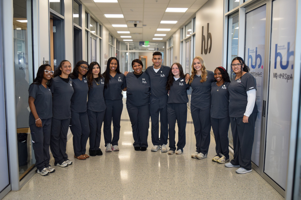 A group of students in uniforms stands in a hallway with glass doors, likely in a medical setting.