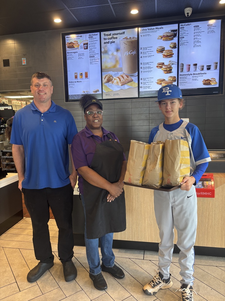 Three people in a fast-food restaurant, one holding two bags of food, stand in front of a menu board.