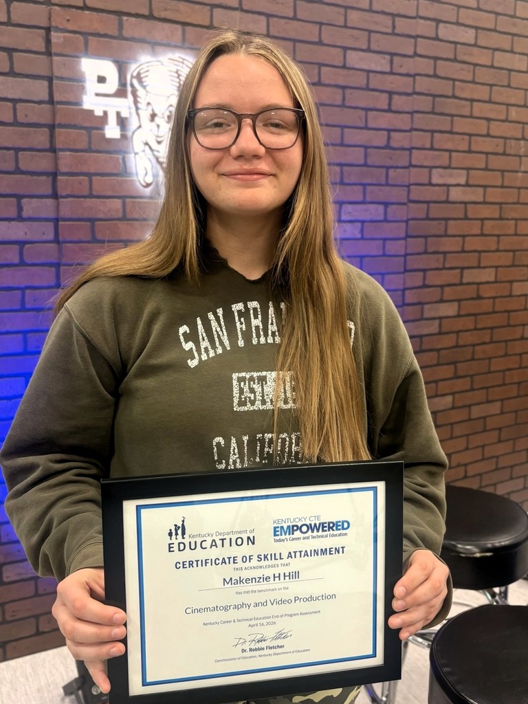 Woman with glasses, a green sweatshirt, and long blonde hair holds a framed certificate. Background includes a brick wall and a sign.