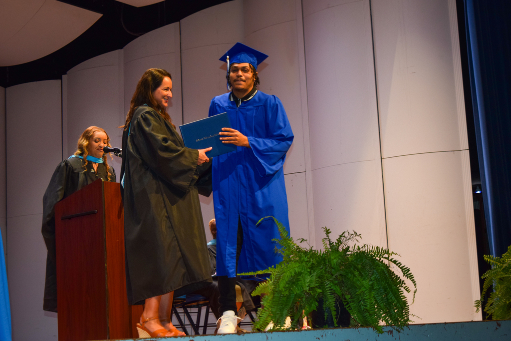 Two individuals in graduation robes, one holding a diploma, stand on a stage. A woman stands behind a podium.