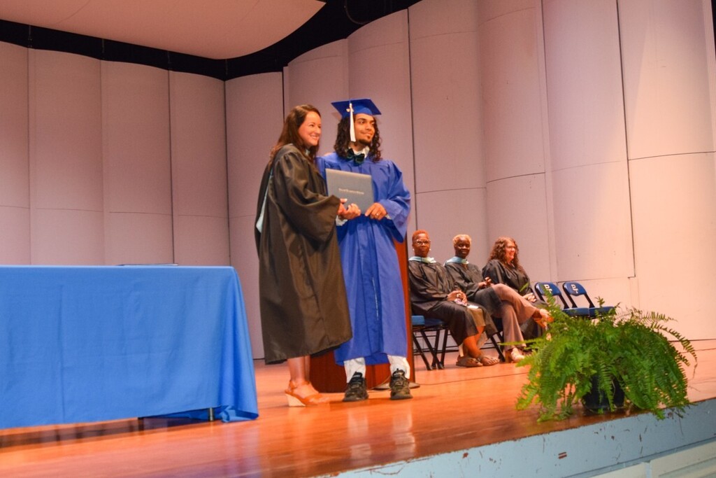 A man in a blue gown receiving a diploma on a stage. Another woman stands beside her. Some audience members sit behind them.