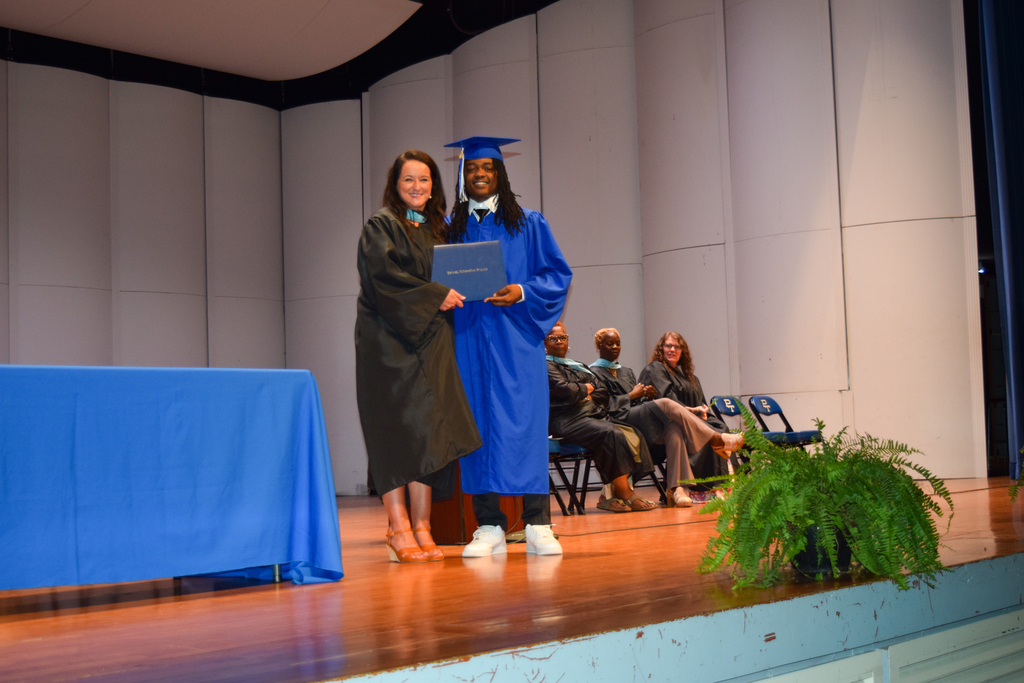 A person in a blue gown and cap hands a diploma to a woman in a black gown.