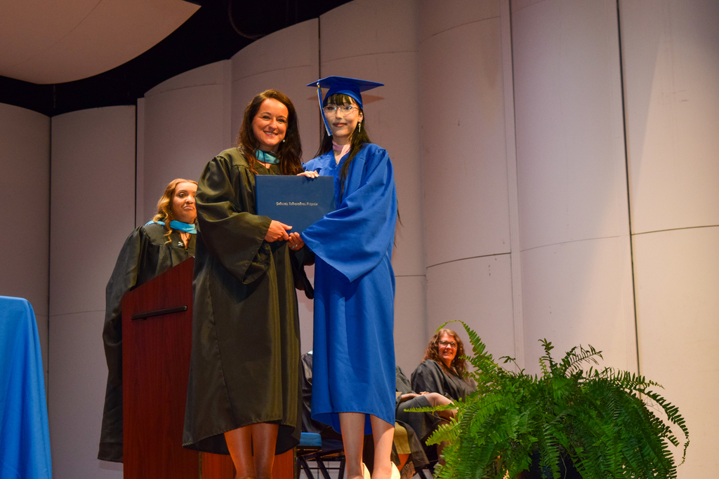 A graduate wearing a blue gown receives a diploma from a faculty member on a stage.