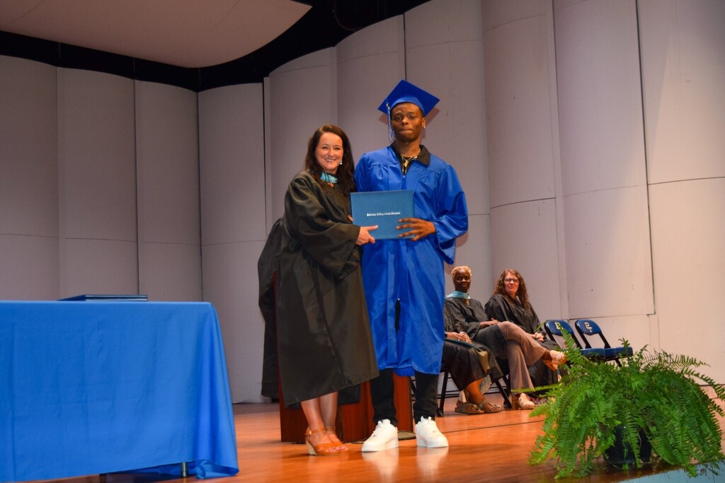 A woman presents a blue diploma to a man wearing a cap and gown on a stage.
