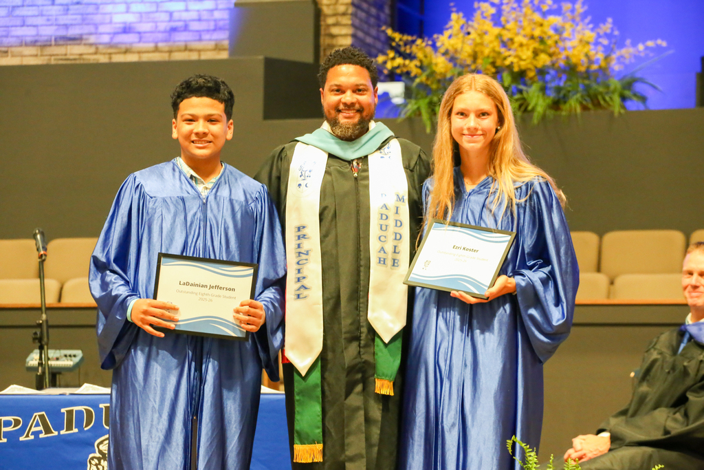 Three people stand on stage in graduation attire, each holding a certificate. The middle person wears a graduation gown and stole.