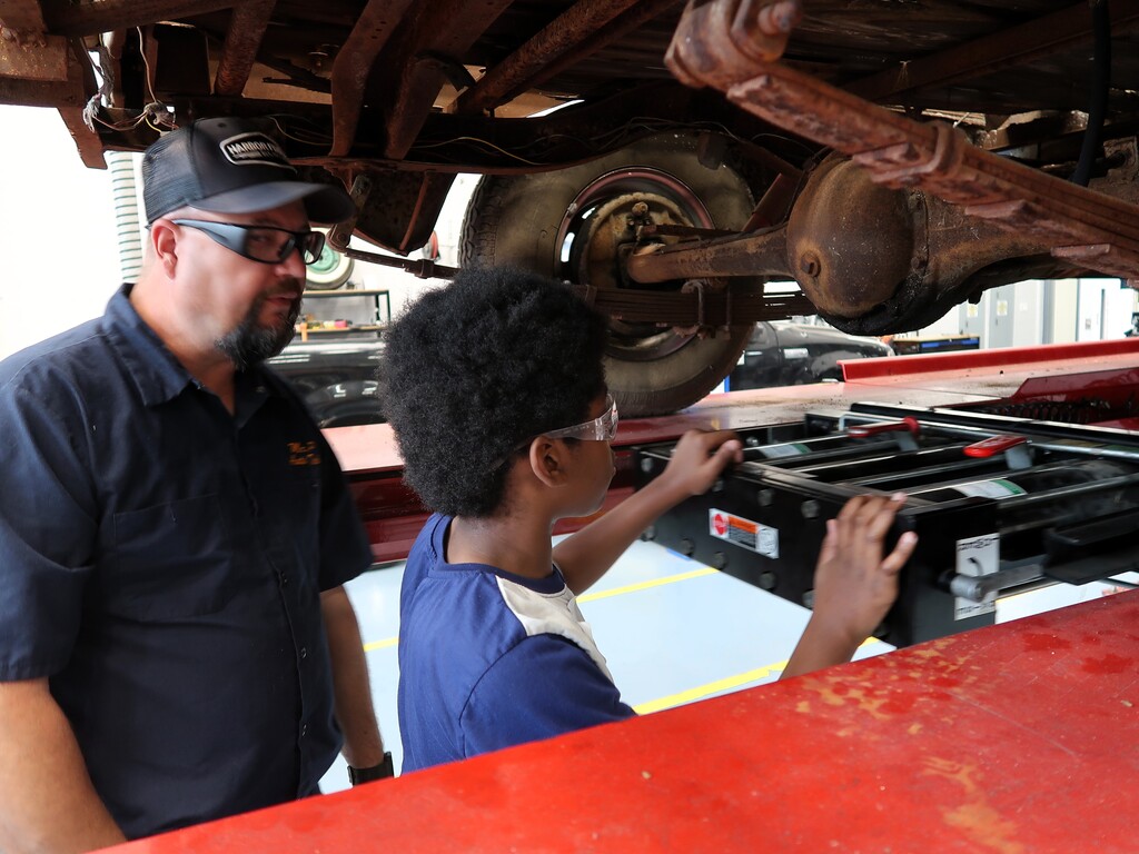 A man and a boy examine the underside of a vehicle. The man wears a cap and glasses.