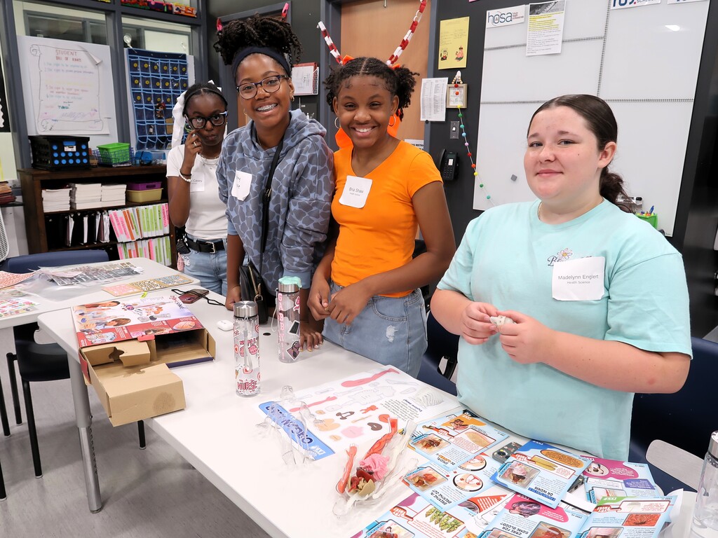 Four women gather at a table, smiling. Various items are on the table, including a box, a water bottle, and colorful materials.