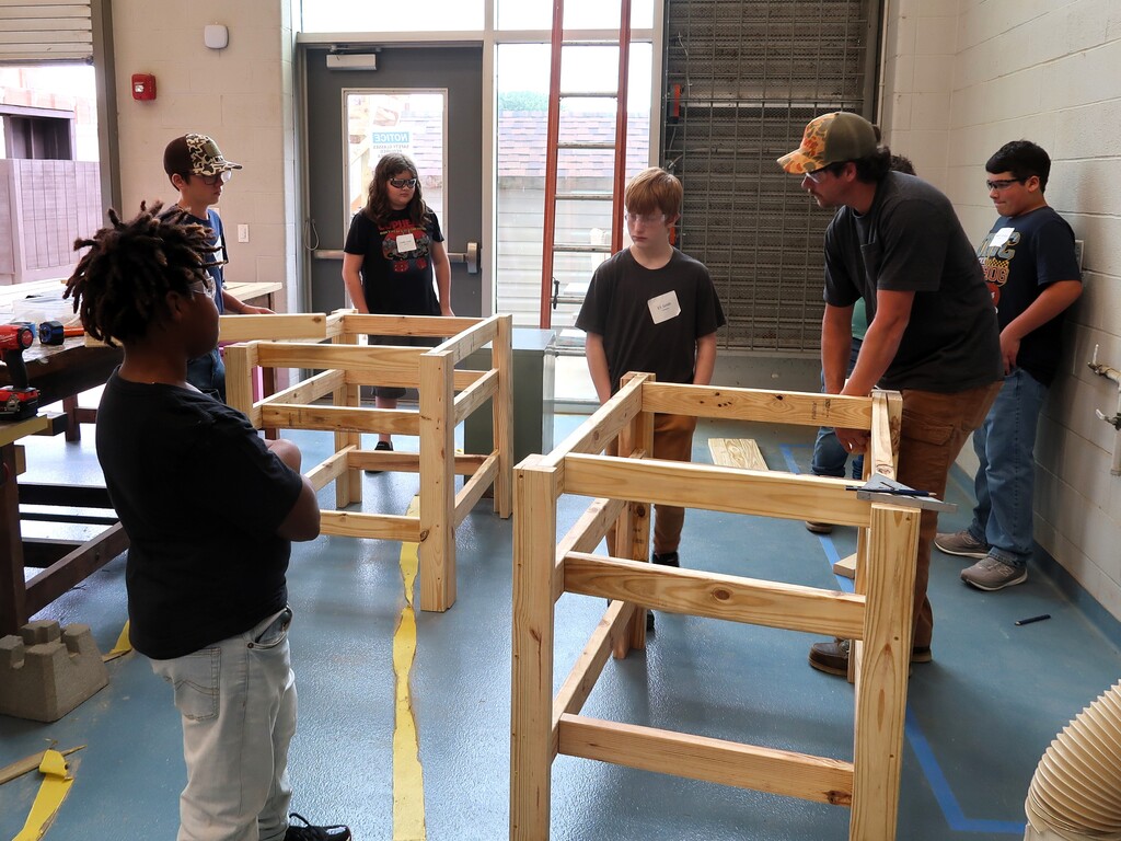 Several individuals in a workshop, including youth, are gathered around wooden structures, with one person in a cap holding a tool.