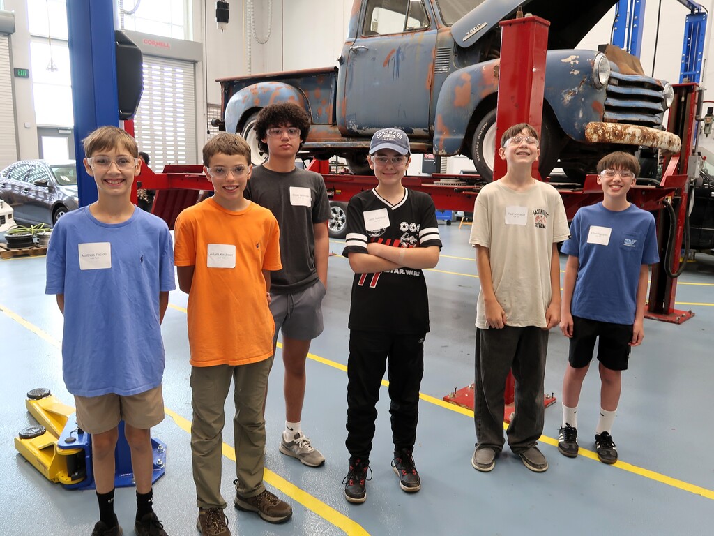 Six boys in a car repair shop. One holds a car jack. An old car on a lift is behind them.