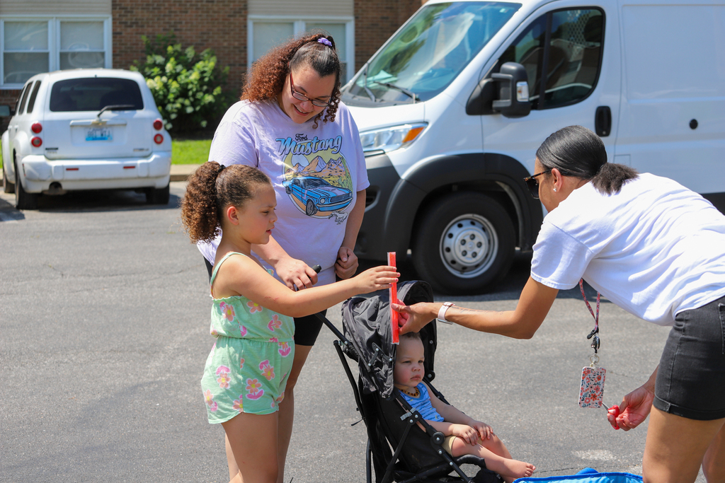 Three people outside with a van in the background, one hands a child a red object.