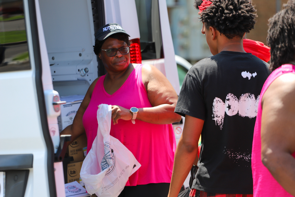 A woman in a pink tank top retrieves items from a van. Two men stand nearby, one in black, the other in pink.