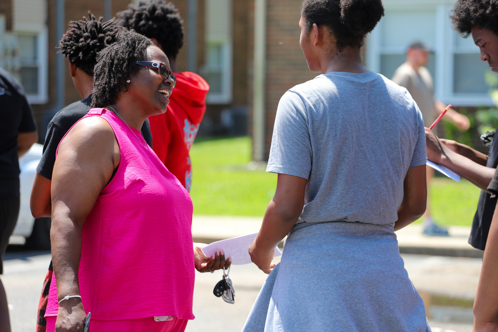 A group of people stand in a street; a woman in a pink top speaks with a woman in a gray dress.