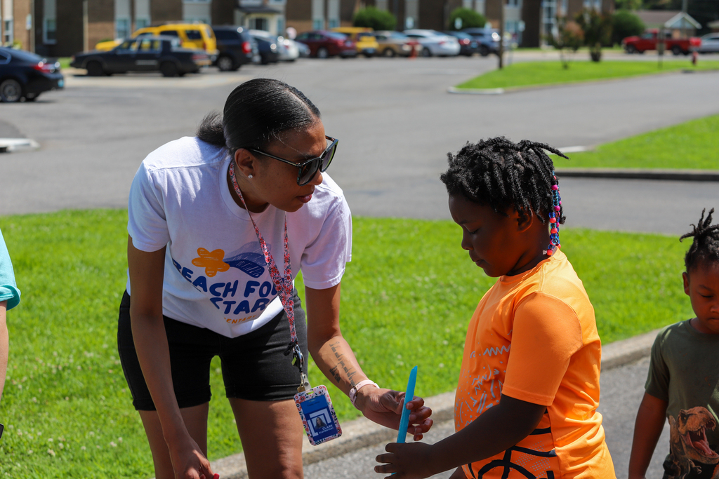 A woman hands a child a toothbrush. Another child stands behind them. A parking lot with cars is in the background.