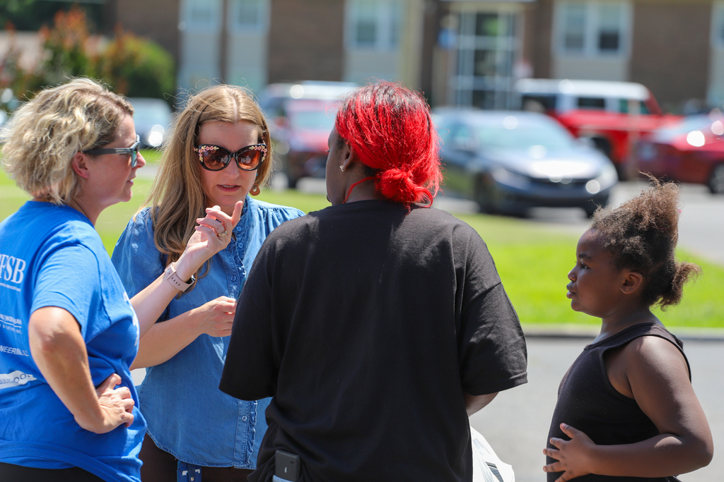 Three women and a girl stand in a parking lot. One woman has red hair, one wears sunglasses, another wears a watch.