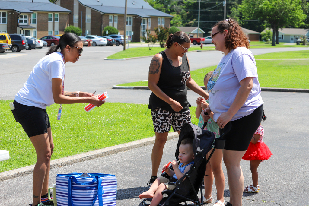 Three women and two children interact near a curb; one woman holds a baby in a stroller.