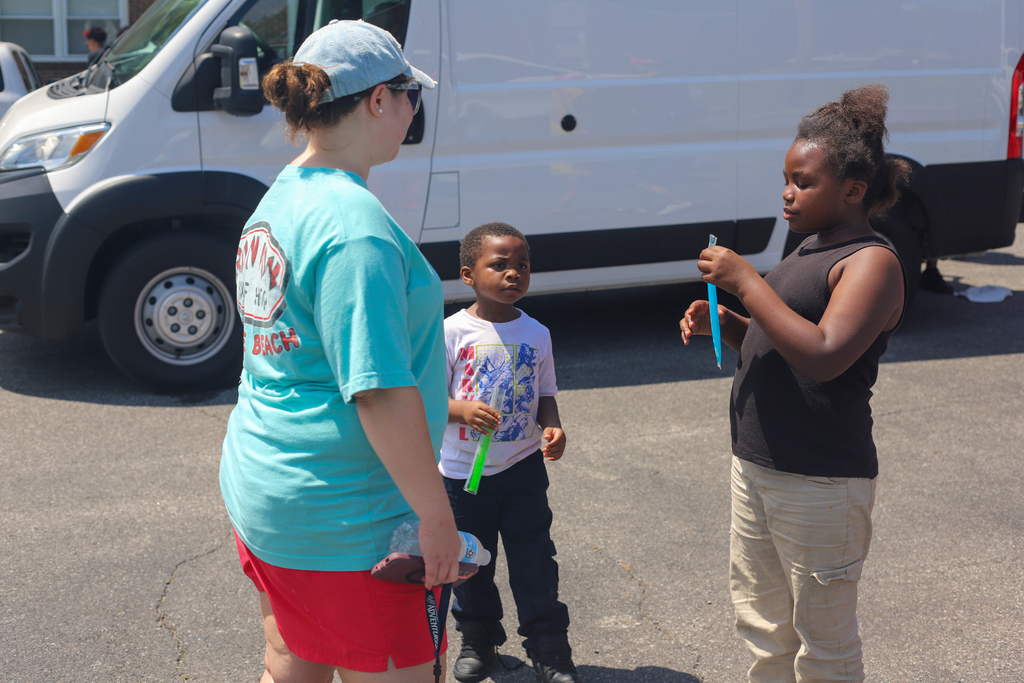 Adult and two children in a parking lot, one holding toothbrushes. White van behind them.