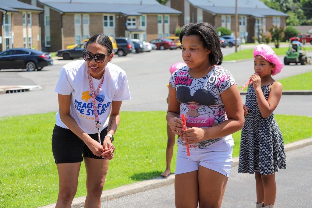 Three girls stand on the street, with two holding toys and the third looking down. Behind them, grass and parked cars surround an apartment complex.