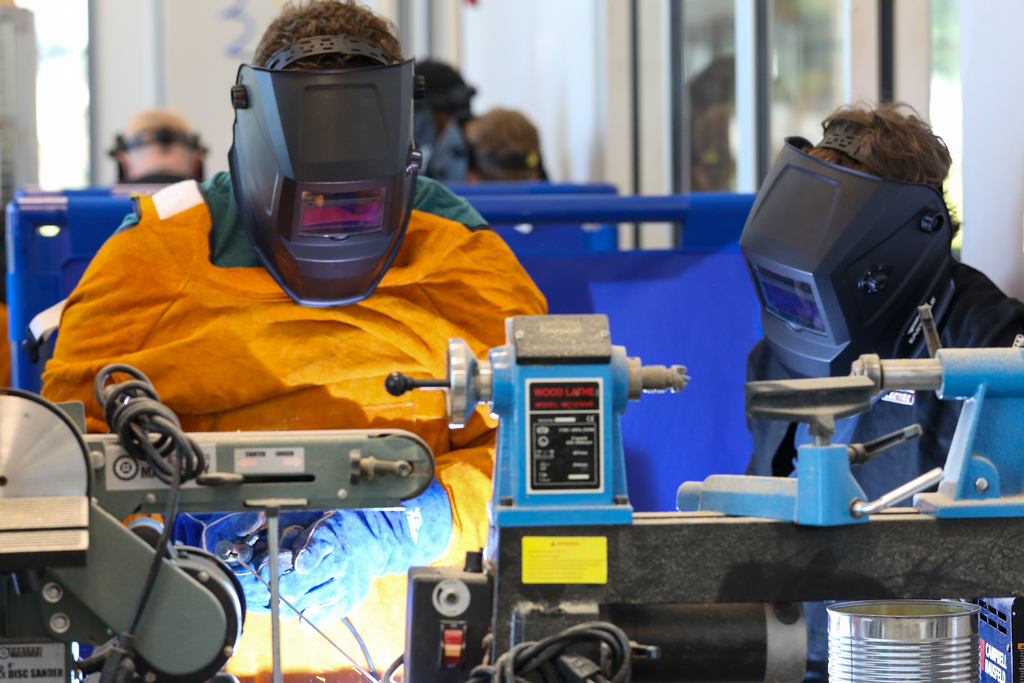 Two individuals wear welding masks, working on metal. One stands in front of a lathe machine.