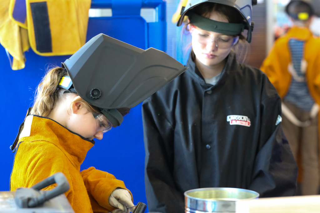 Two students wearing safety gear in a workshop. One has a welding mask on, and the other stands nearby.