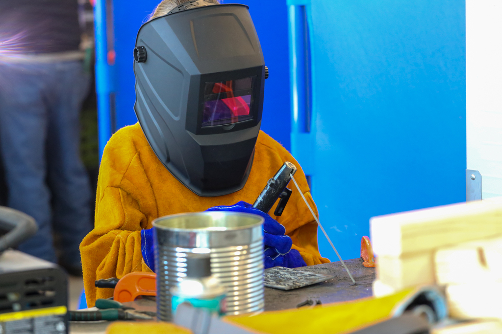 Person in welding gear with a mask on, holding a welding tool, sits at a table with a tin can.