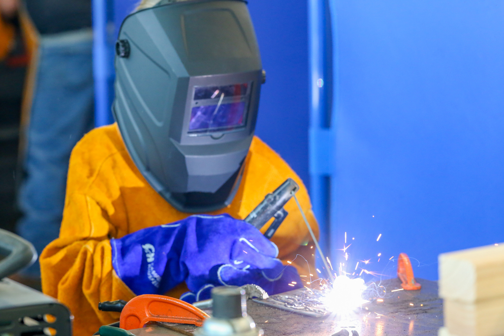 Person in yellow protective clothing and mask welding at a workstation with sparks. Blue background.