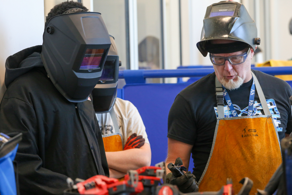 Two men wearing safety gear, one in a welding mask, in a workshop. One man has an apron.