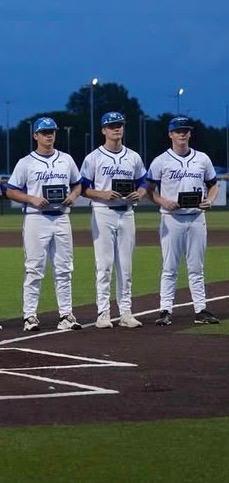 Three players in matching jerseys stand on a baseball field at night. Each holds a trophy.