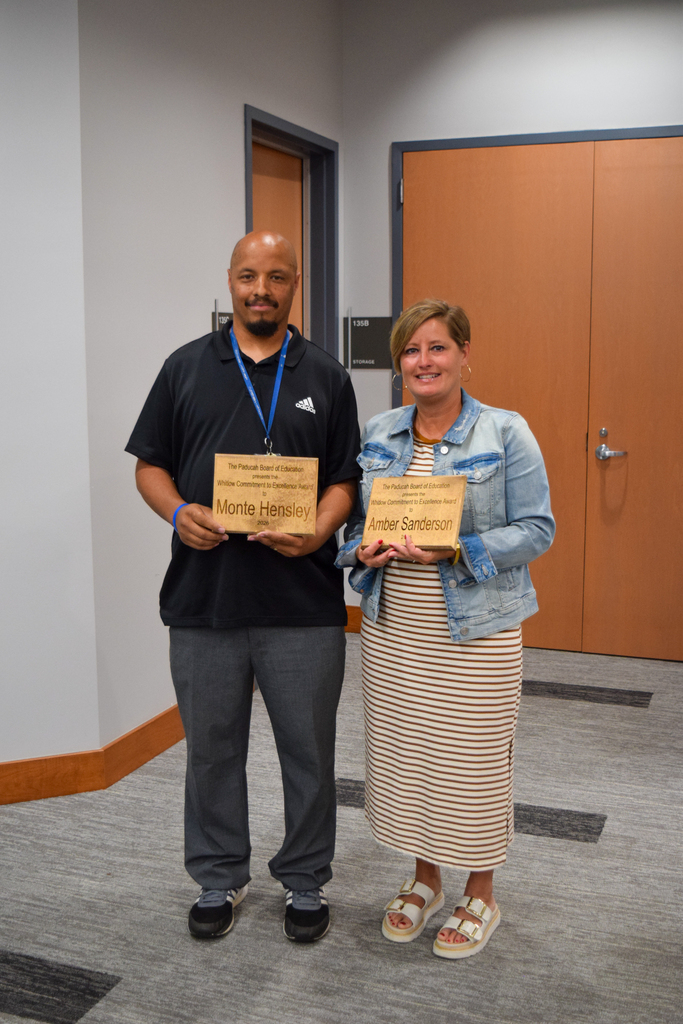 Two people stand in a room with wooden doors, holding awards. One person wears a black shirt and gray pants, and the other a denim jacket and striped skirt.