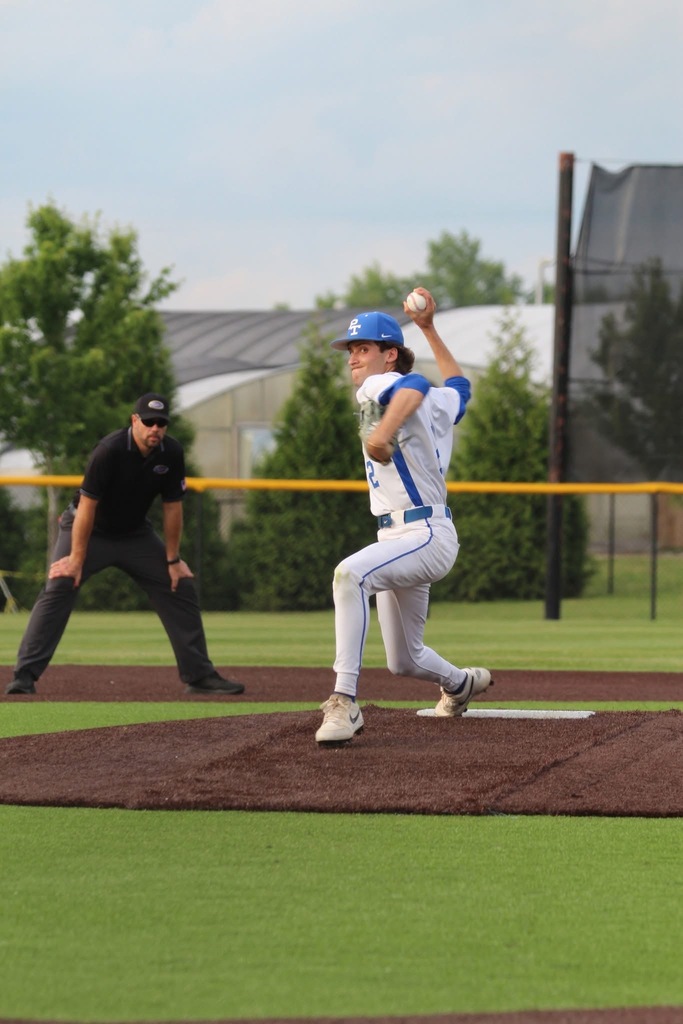 Baseball player in blue and white uniform pitches from the mound; an umpire watches from behind, with a green fence and trees in the background.