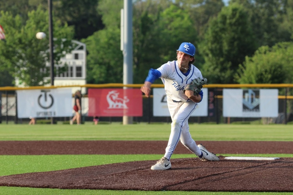 Baseball player in blue hat and uniform throwing a ball on a baseball field with banners in the background.