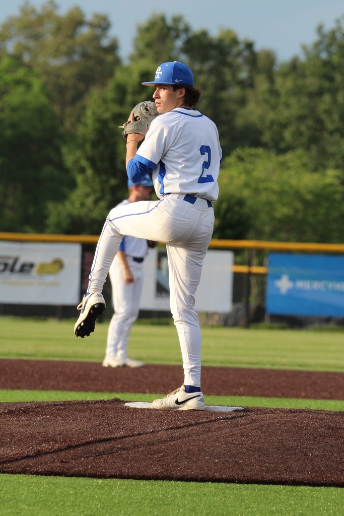A baseball player in a blue cap winds up for a pitch on a baseball field. Behind them is a blurred background with banners and trees.