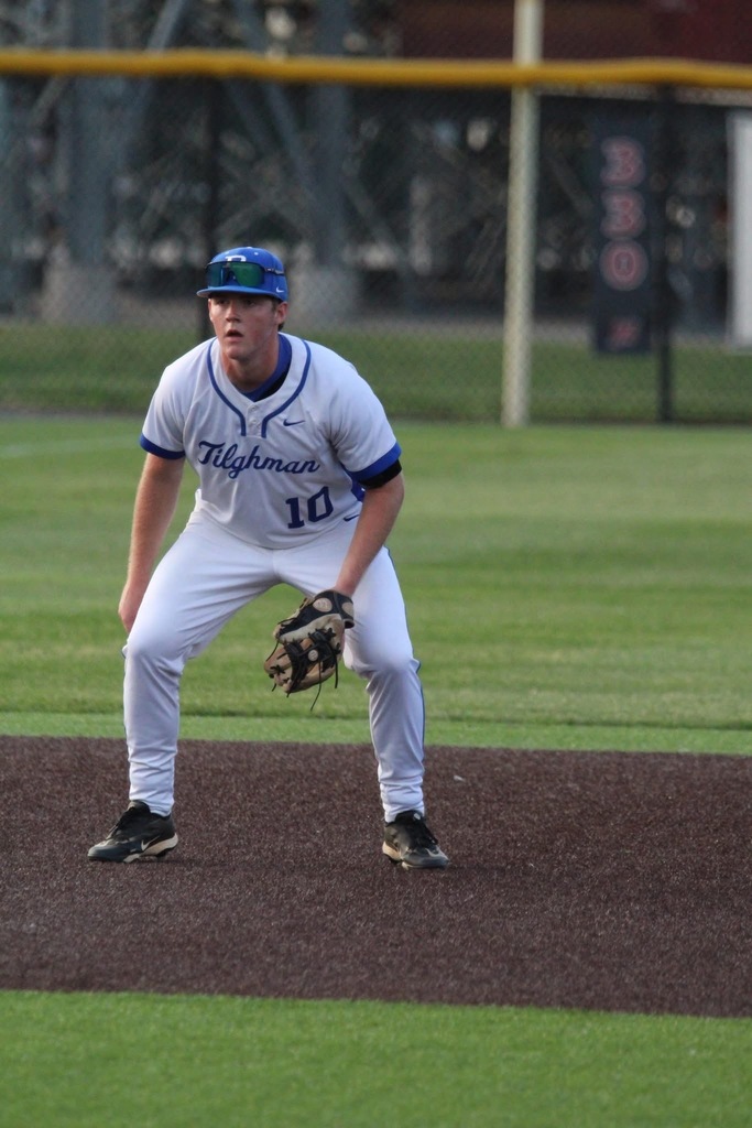 A baseball player in white and blue stands on a field. The player wears a blue cap, a baseball glove, and number 10.