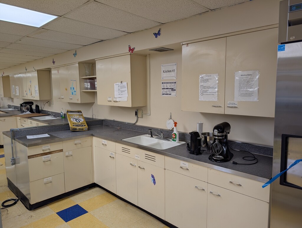 A kitchen with white cabinets, countertops, and appliances. Tiled floor with blue markers. Overhead lighting.