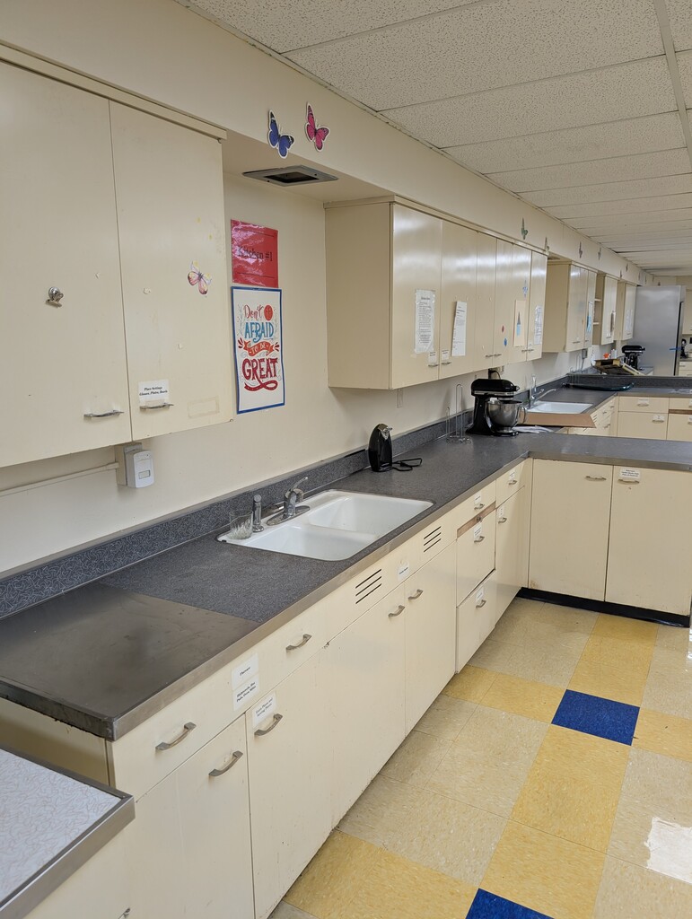An empty kitchen with cabinets, a countertop, sink, and checkered floor. A refrigerator is visible at the far end.