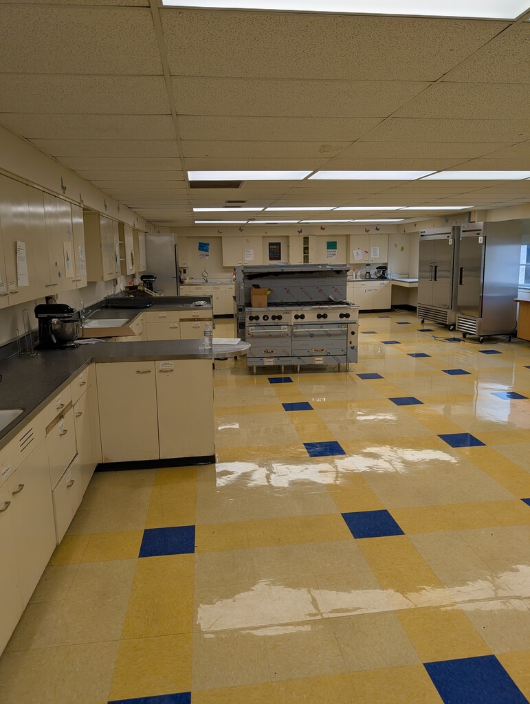 A commercial kitchen with tiled floor, white cabinets, and stainless steel appliances. Fluorescent lights on the ceiling.