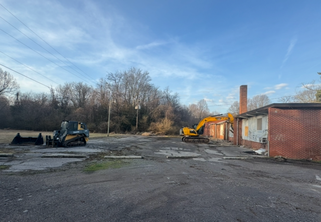 A construction site with a yellow excavator and a large, open area. A brick building with a chimney.