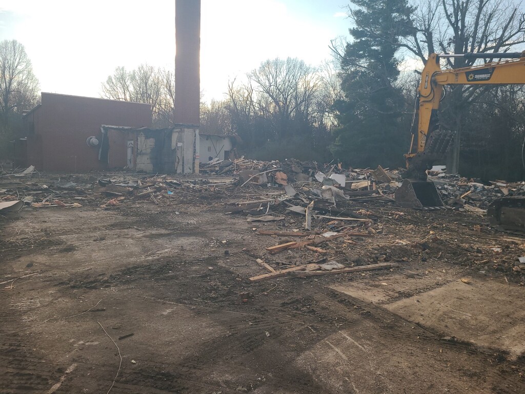 Construction site with debris and a yellow backhoe in front of a building with a tall chimney.