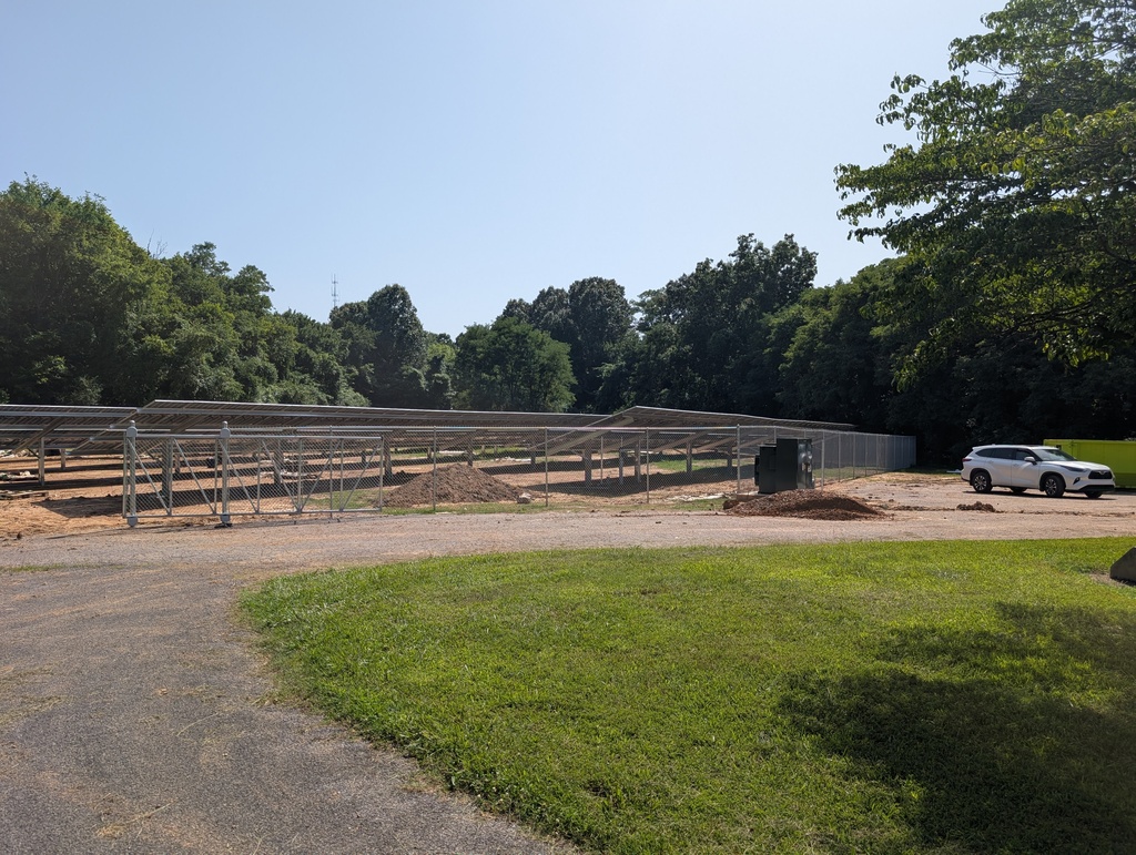 A solar farm under construction. A car is parked in front of the fence. Green grass and trees are in the background.