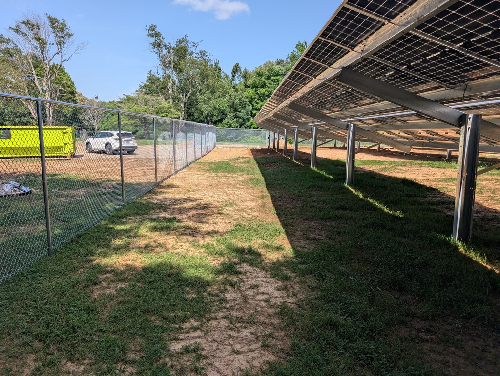 Solar panels are installed on a metal structure next to a fenced yard with a car parked in the background.