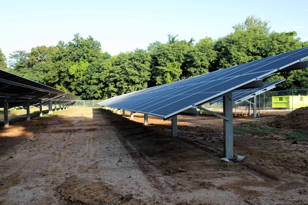 Solar panels installed on metal stands in a dirt field, surrounded by trees and greenery.