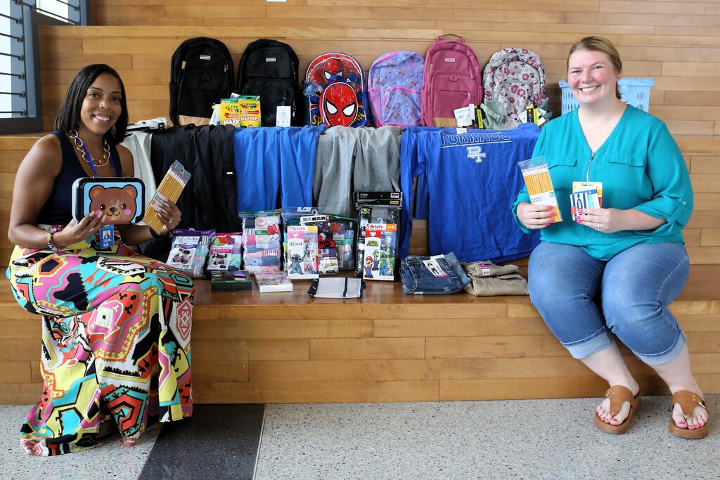 Two women sit on a bench holding items. Behind them, a table holds backpacks, clothing, and other items.