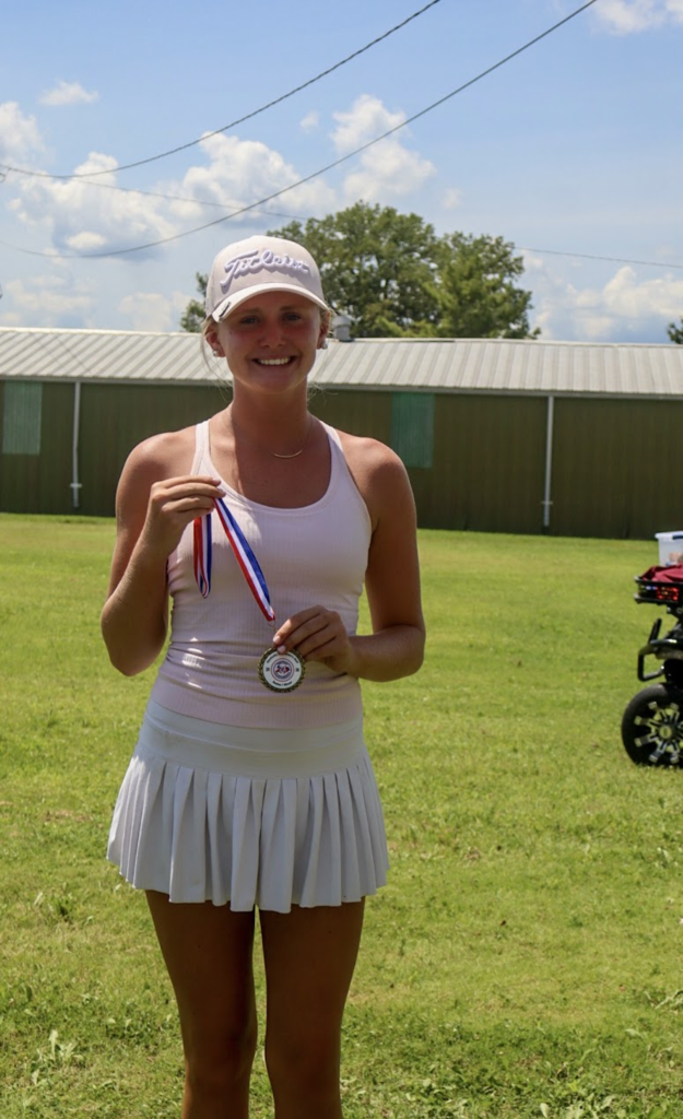 A woman in a white tennis outfit holds a medal with a ribbon around her neck. Behind her is a grassy area and a building.