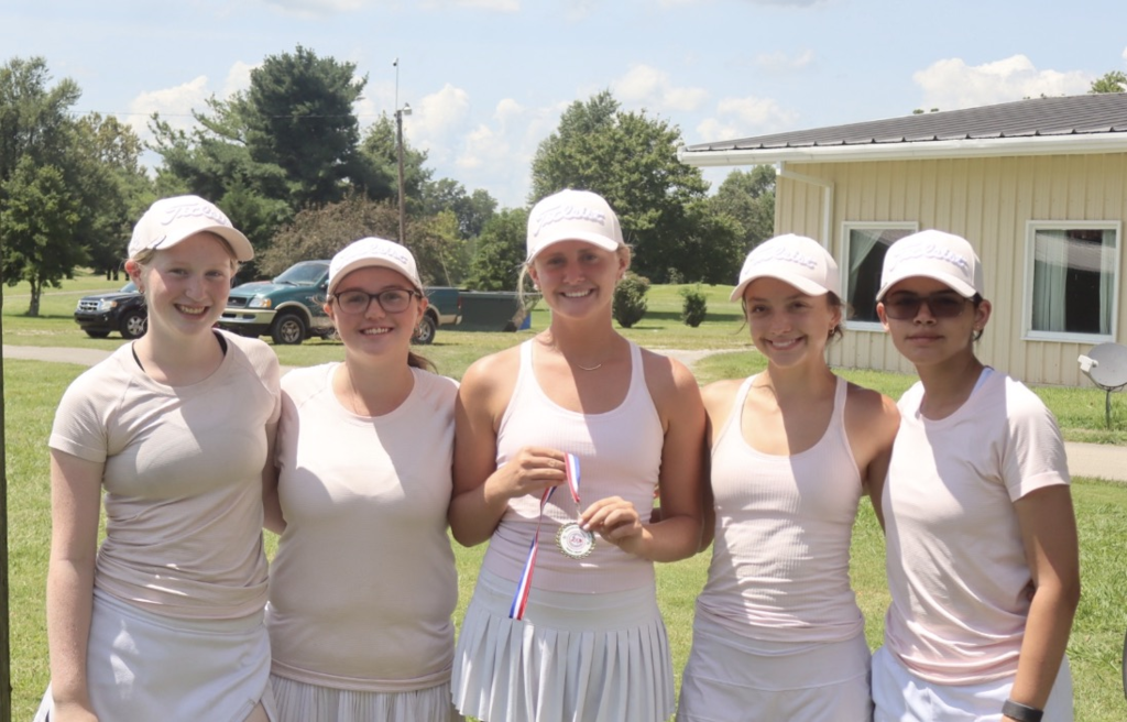 Five women in white tennis attire and caps pose with medals behind them in a grassy field.