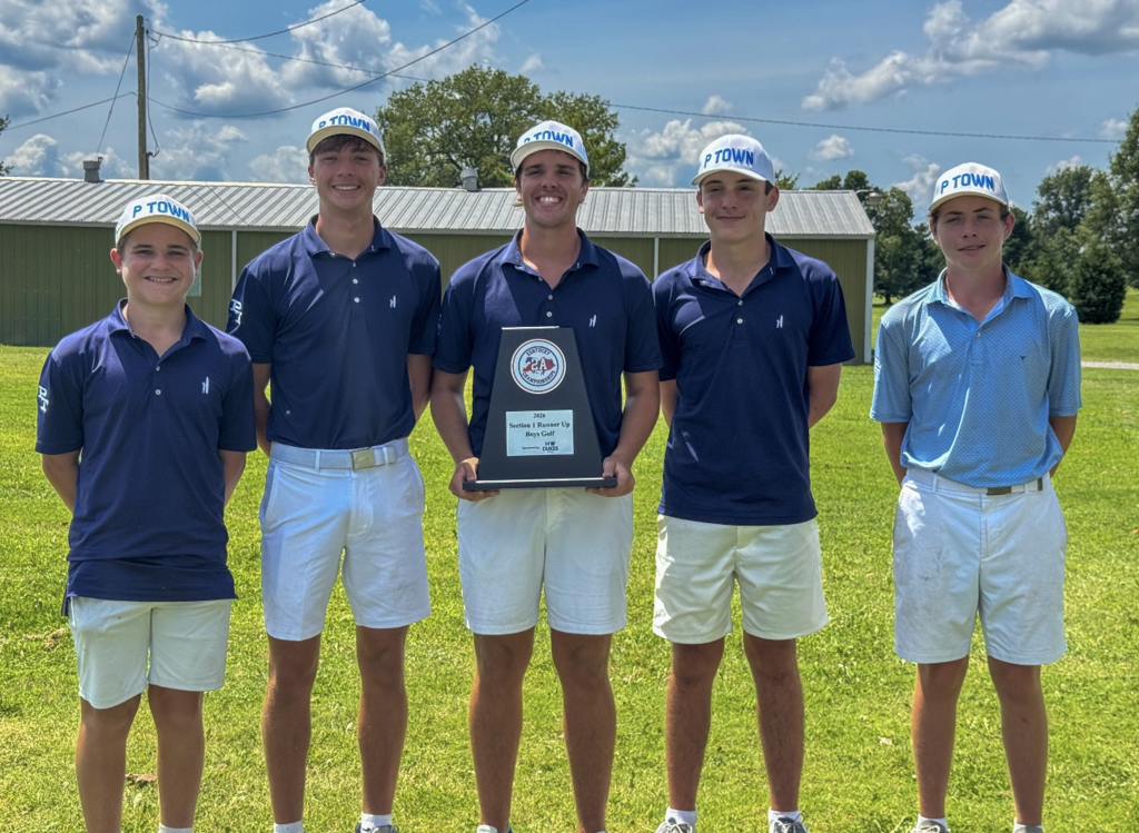 Five young men in matching blue shirts and white shorts stand in a grassy field with a trophy.
