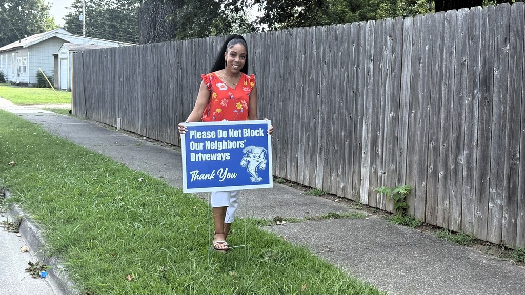Woman holding a blue sign that says "Please do not block for neighbors driveways" in front of a wooden fence.