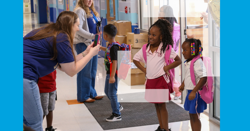 A woman in blue holds a phone near two young girls with backpacks. Nearby, another child stands with a backpack.