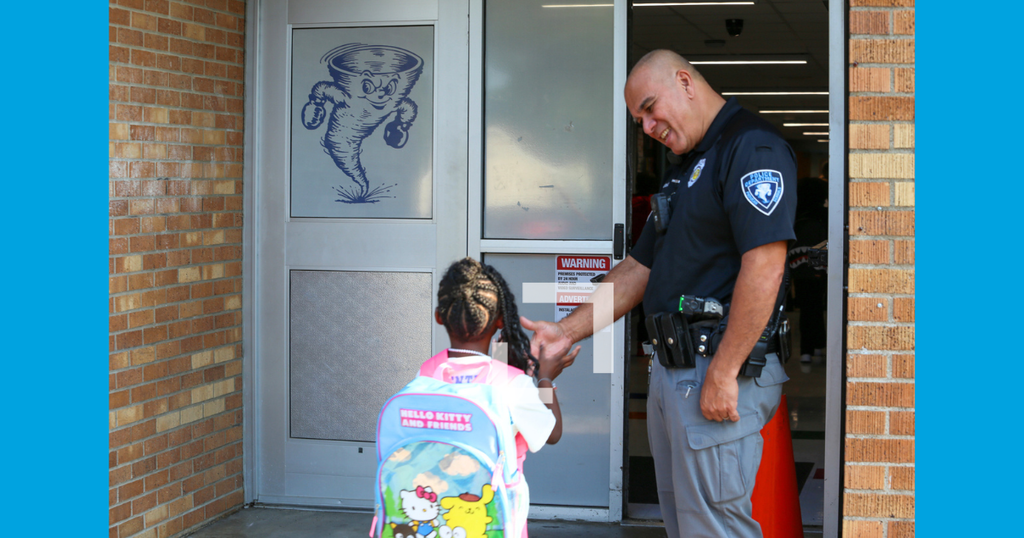 A police officer is greeting a young child in a hallway, with a brick wall and blue background.