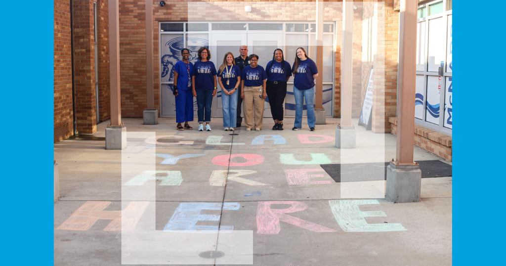 A group of people in matching shirts stand near a building. The floor has colorful chalk writing.