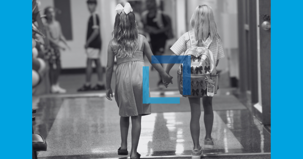 A black-and-white photo of two children holding hands in a hallway. The scene also shows other children walking in the background.